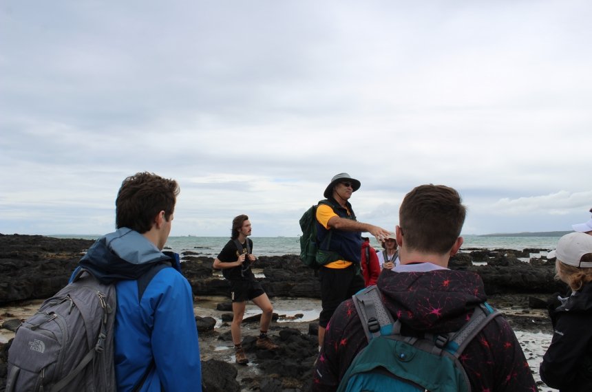 A professor addresses students gathered on a block rocky beach created by lava flowing into the ocean.