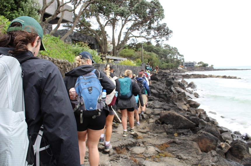 A line of students walk along a black rocky coastline.