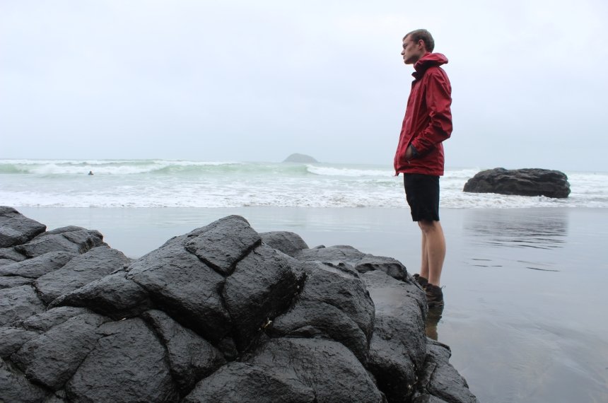 A man stands on a beach alongside volcanic rocks as waves crash behind him.