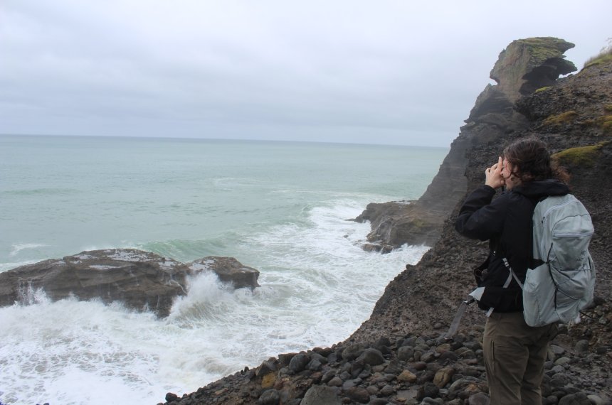 A woman looks off a rugged rocky cliff as ocean waves crash beneath her.