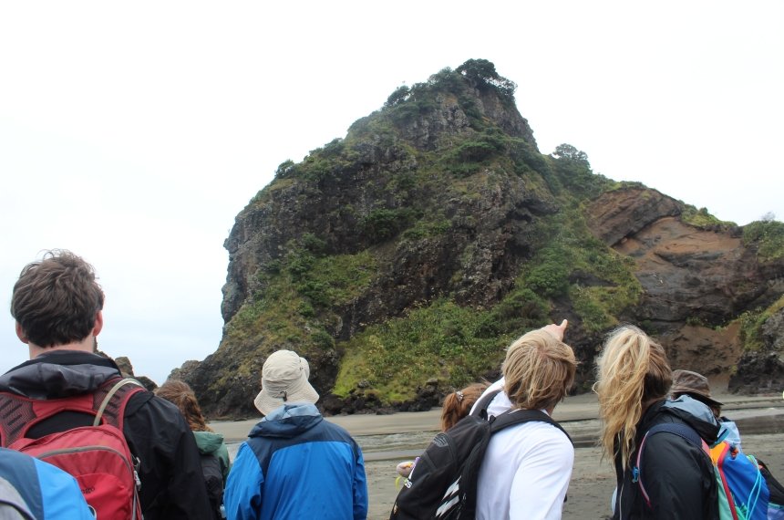 Students point and look at a large rock outcropping rising from a sandy beach.