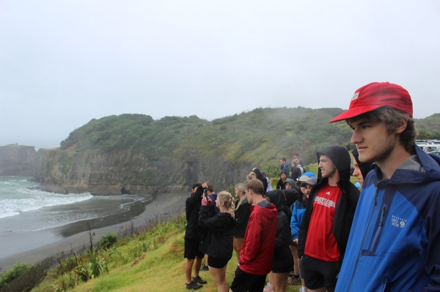 Students stand on a hillside looking at the ocean with a rugged coastline in the background.