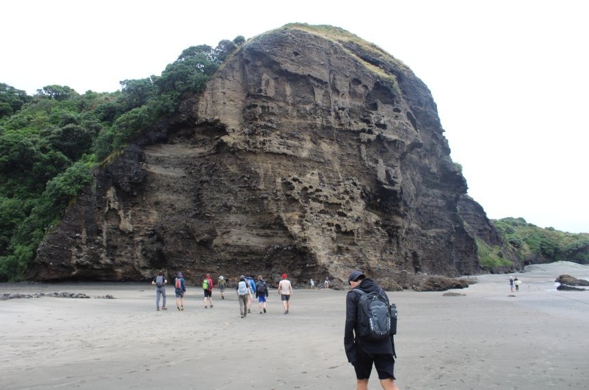 Students walk across a sandy beach toward a large cliff face.