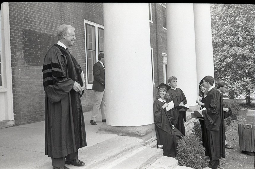 Jimmy Carter stands on the steps of Old Centre wearing black graduation robes as school officials watch.