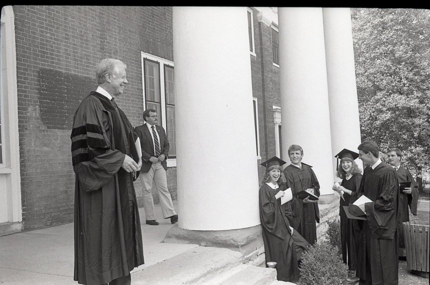 Jimmy Carter stands on the steps of Old Centre wearing black graduation robes as school officials watch.