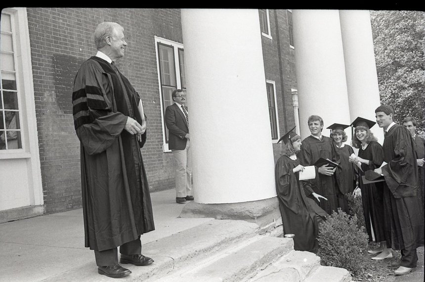 Jimmy Carter stands on the steps of Old Centre wearing black graduation robes as school officials watch.