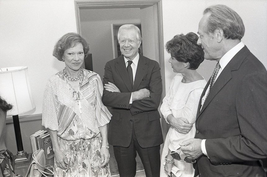 Former President Jimmy Carter stands with his arms crossed as he speaks with two women and a man.