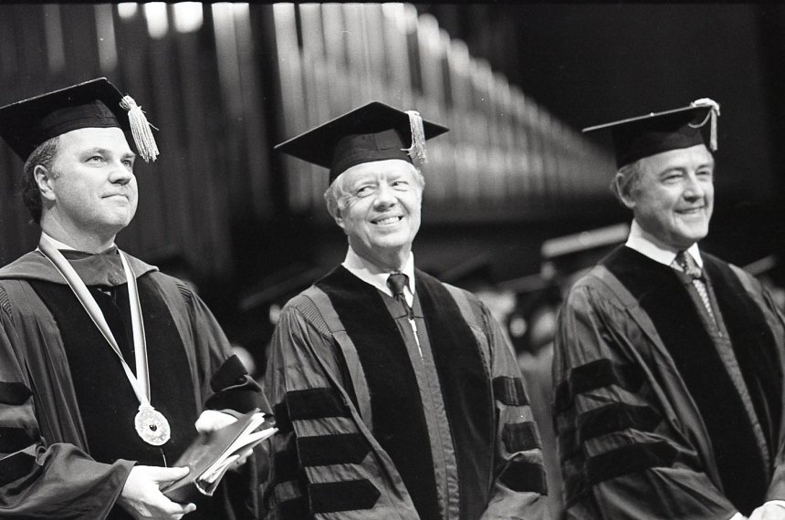 Former President Jimmy Carter wears a graduation robe and cap while standing between who others during commencement.