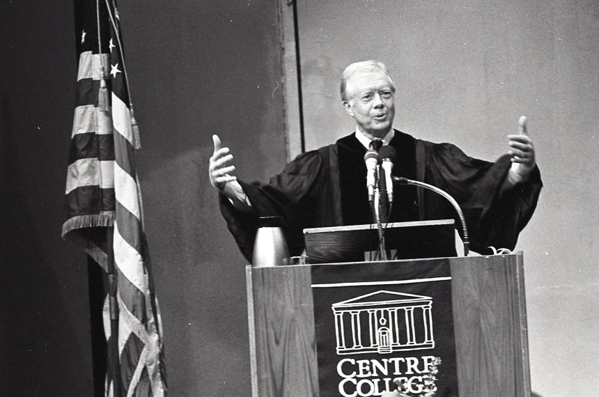 Former President Jimmy Carter spreads his arms while speaking behind a podium bearing the Centre College name and logo.