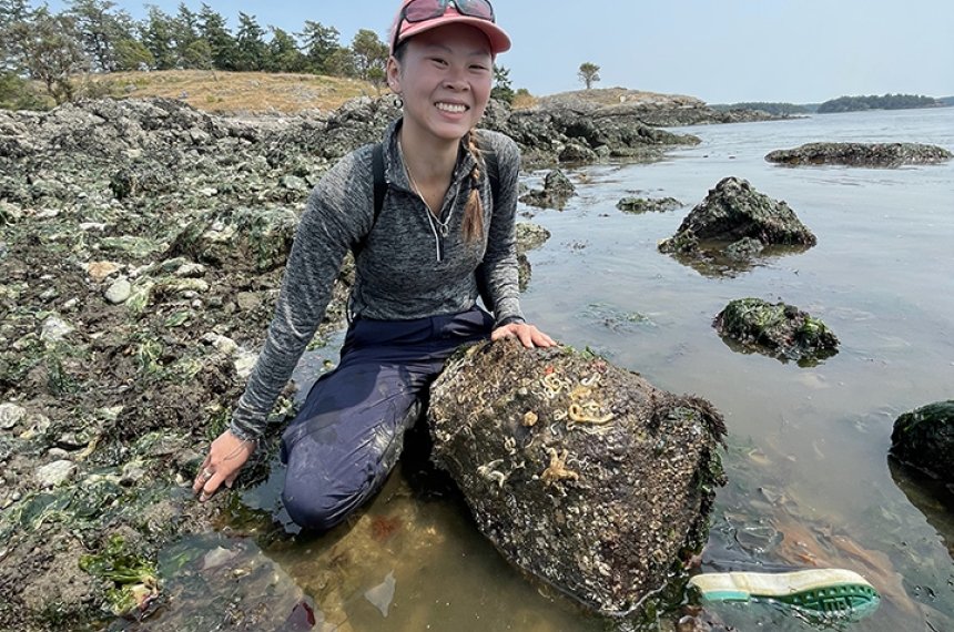 A young woman kneels behind a large stone where a rocky beach meets the ocean.