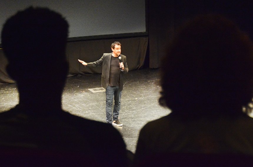 A man holding a microphone is seen between the silhouettes of two audience members.