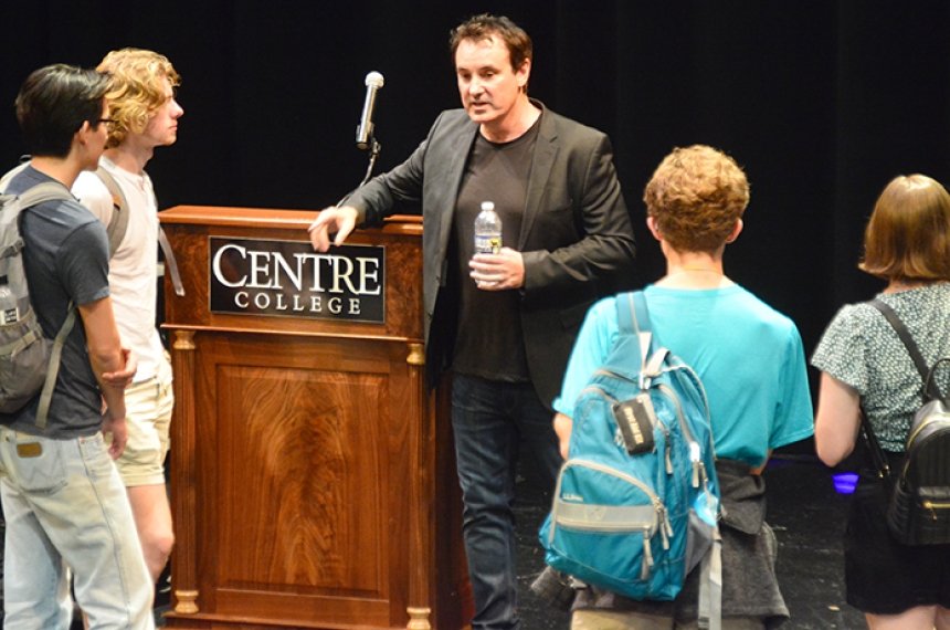 Students carrying backpacks stand in a loose circle around a man holding a water bottle and resting his arm on a podium bearing the Centre College logo.