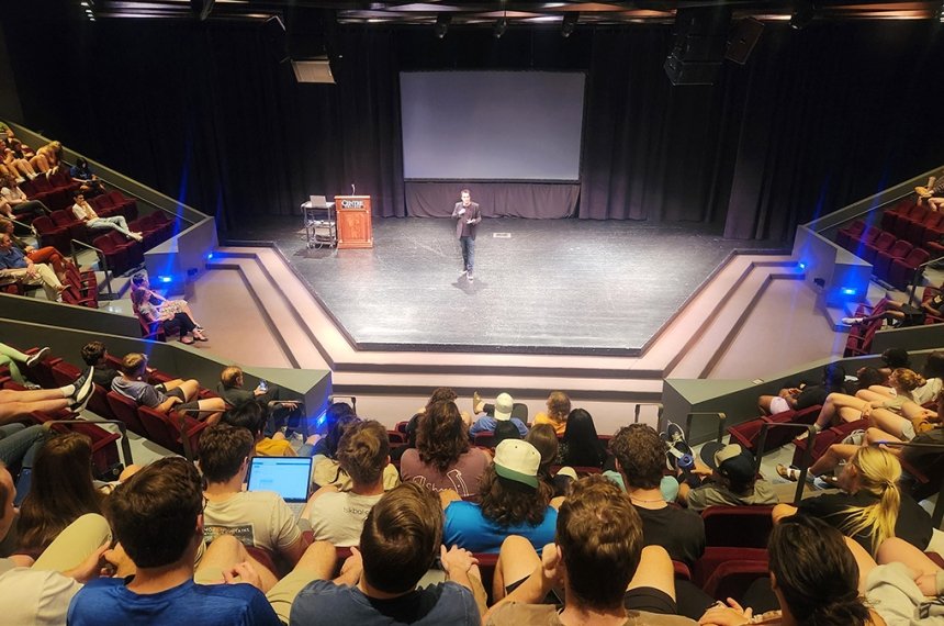 A man talks into a microphone while standing on a stage surrounded by theatre seats filled with students.