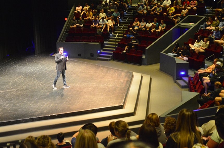 A man talks into a microphone while standing on a stage surrounded by theatre seats filled with students.