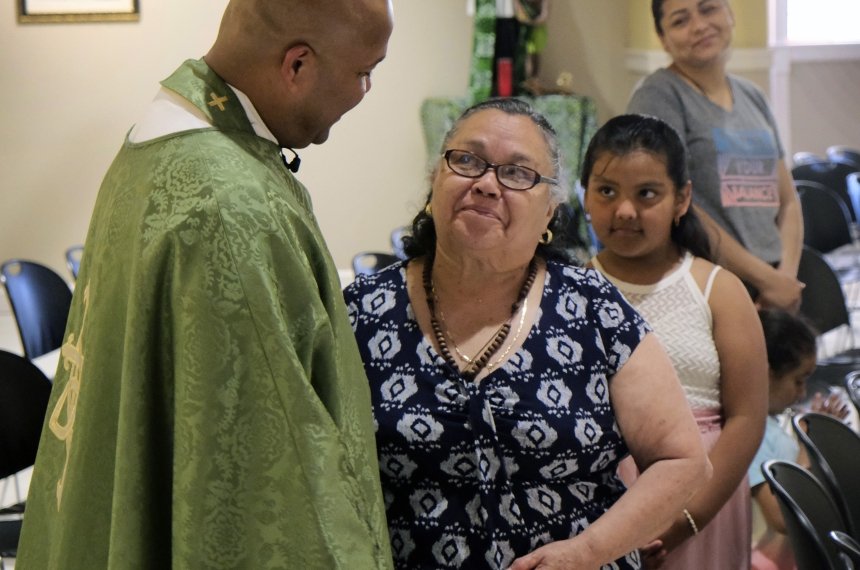 A priest in green and gold robes speaks with a woman as a young girl waits behind her at a religious service.