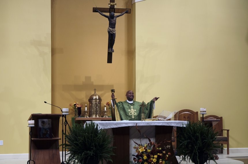 Beneath a crucifix hanging on the ball, a priest wearing green and gold robes holds his hands out during a religious service.