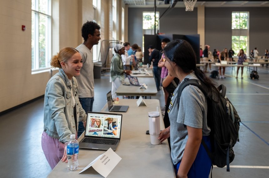 Two young women talk across a folding table while other students chat in the background.