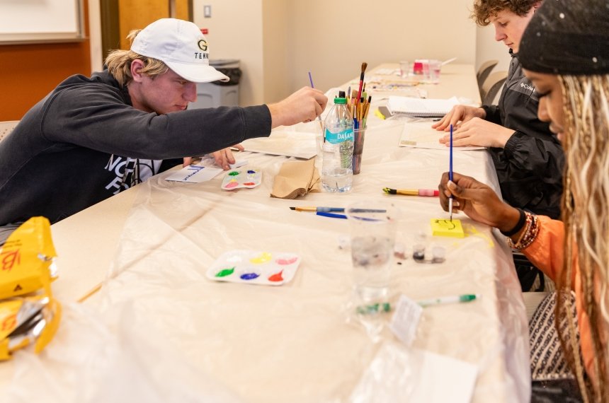 Students seated an opposite sides of a plastic-covered table paint.