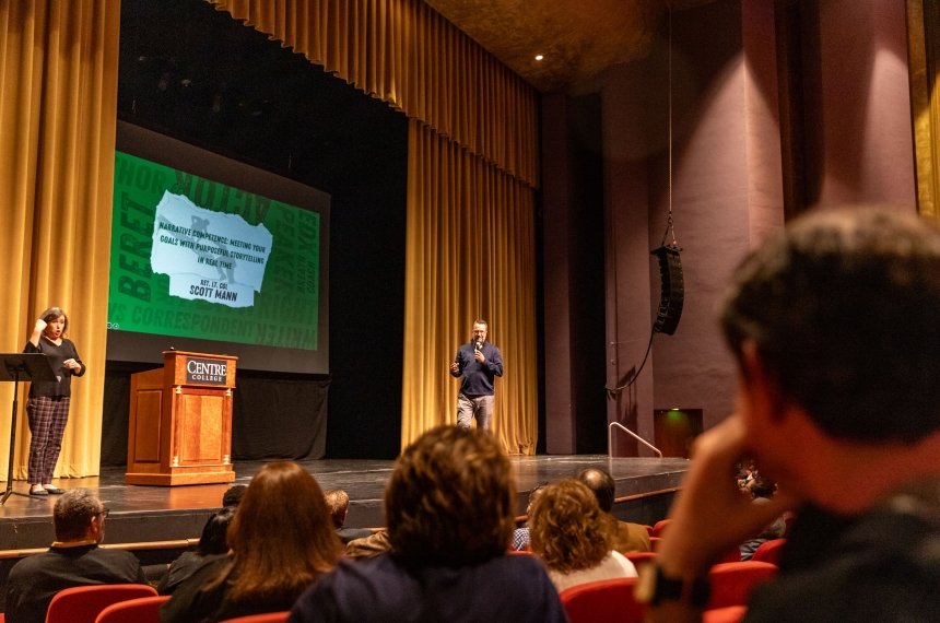 A man holding a microphone on stage speaks to a large audience.
