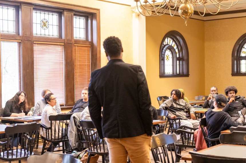 A man stands with his back to the camera as he addresses others seated around tables.