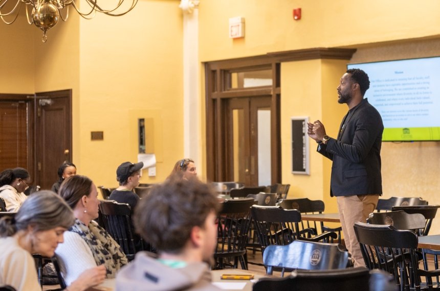 A man stands at the front of a room addresses others seated at tables.