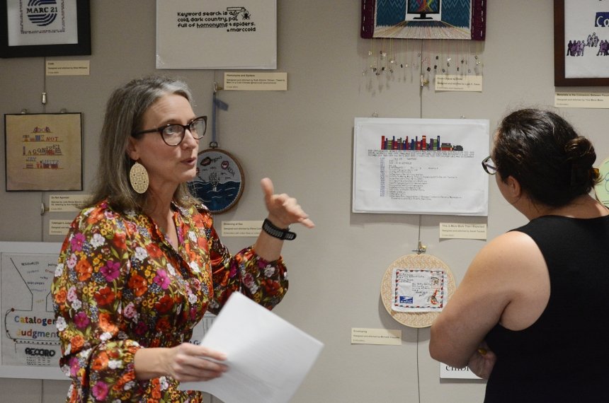 A woman holding papers speaks as another woman looks at textile art displayed on a wall.