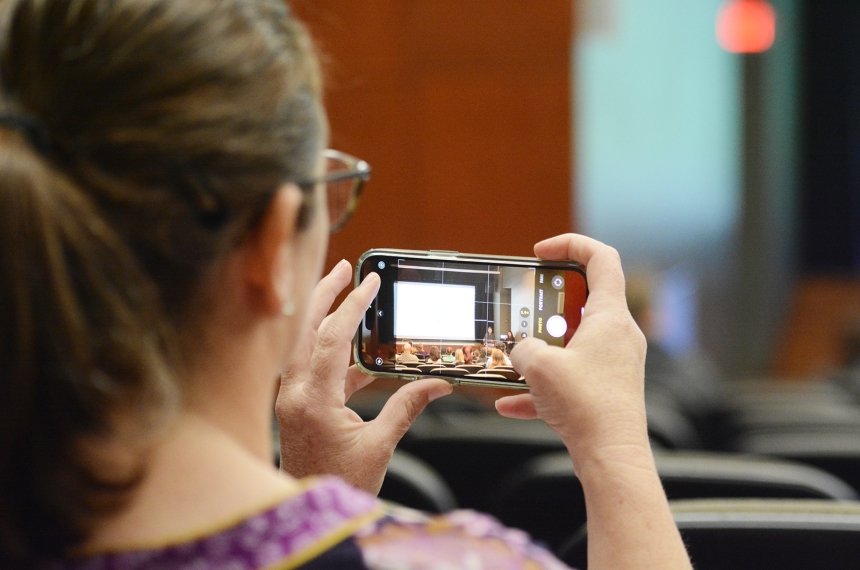 A women uses her cell phone to take photos of a man speaking at the front of a large room.