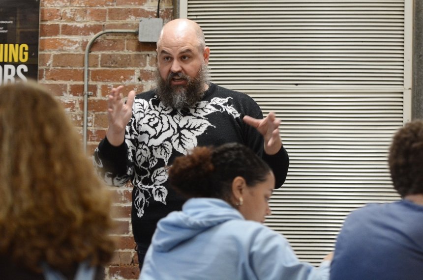 A man addresses students seated at round tables.
