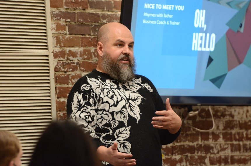 A man stands in front of a screen with the worlds &quot;Oh Hello&quot; on it as she speaks.