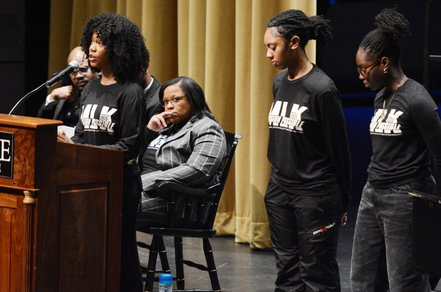 Three Centre College students stand on stage at the Norton Center for the Arts one one speaks into a microphone.