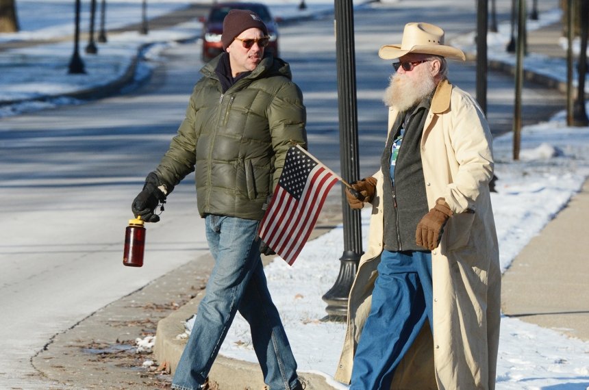 A man holding an American flag marches across the street, accompanied by another man.