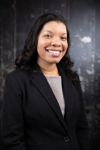 Lady with long dark hair wearing black jacket and grey top in front of black background