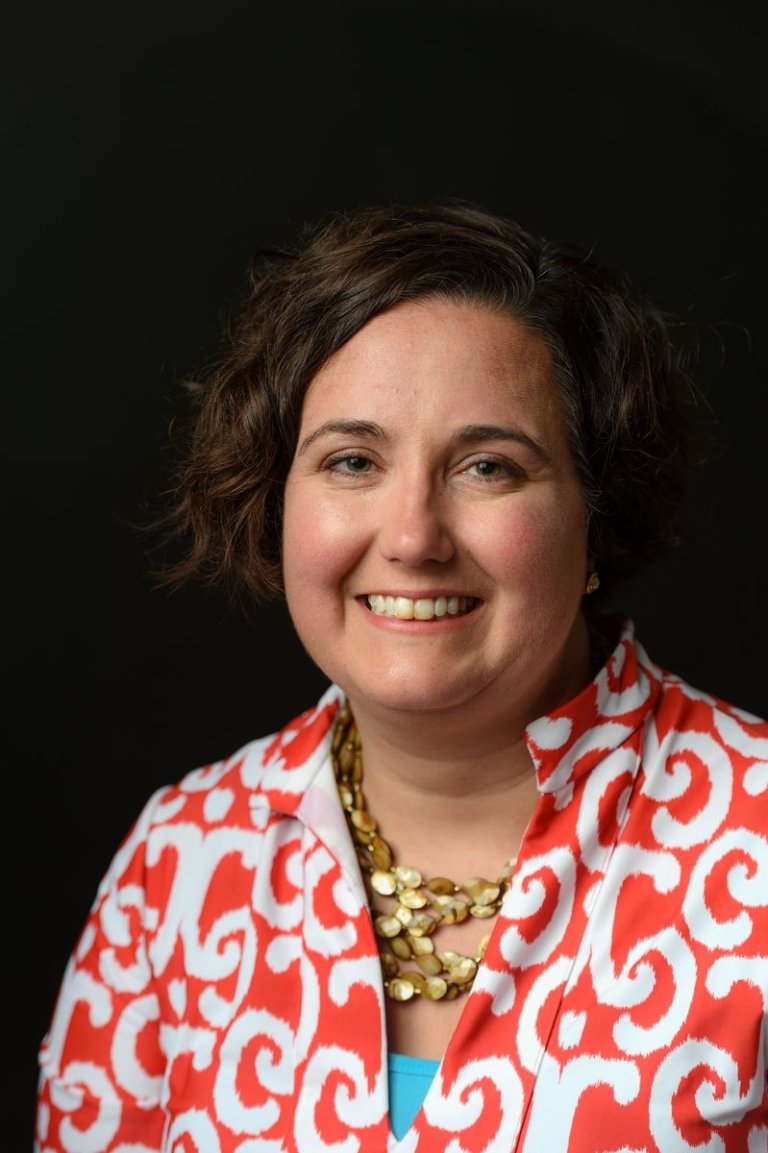 Lady with dark hair wearing red and white patterned top with gold necklace in front of dark background