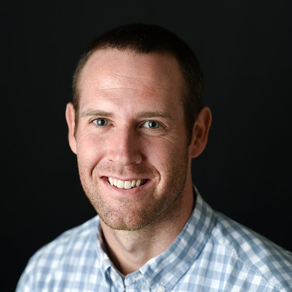 man with short facial hair wearing blue and grey checked shirt in front of dark background