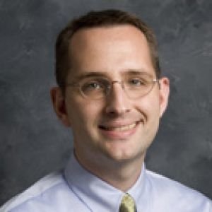 Man with short brown hair wearing glasses, grey button up dress shirt with gold tie in front of grey background