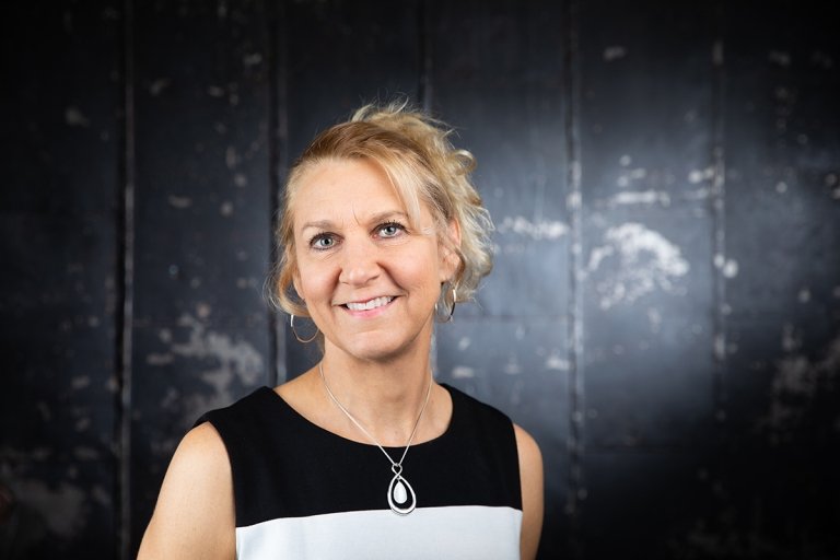 Lady with blonde hair in ponytail wearing black and white top standing in front of black backdrop