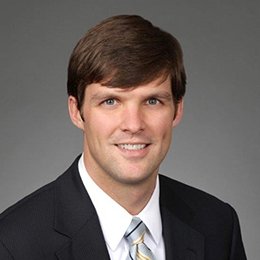 Person standing in front of grey background wearing dark suit