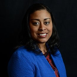 Person wearing royal blue jacket standing in front of dark background smiling
