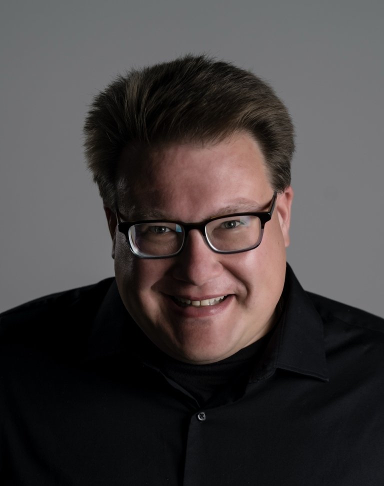 Man with short brown hair wearing glasses and black button up shirt in front of grey background