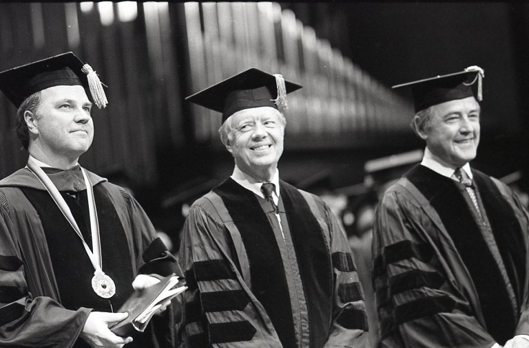 Former President Jimmy Carter wears a graduation robe and cap while standing between who others during commencement. 