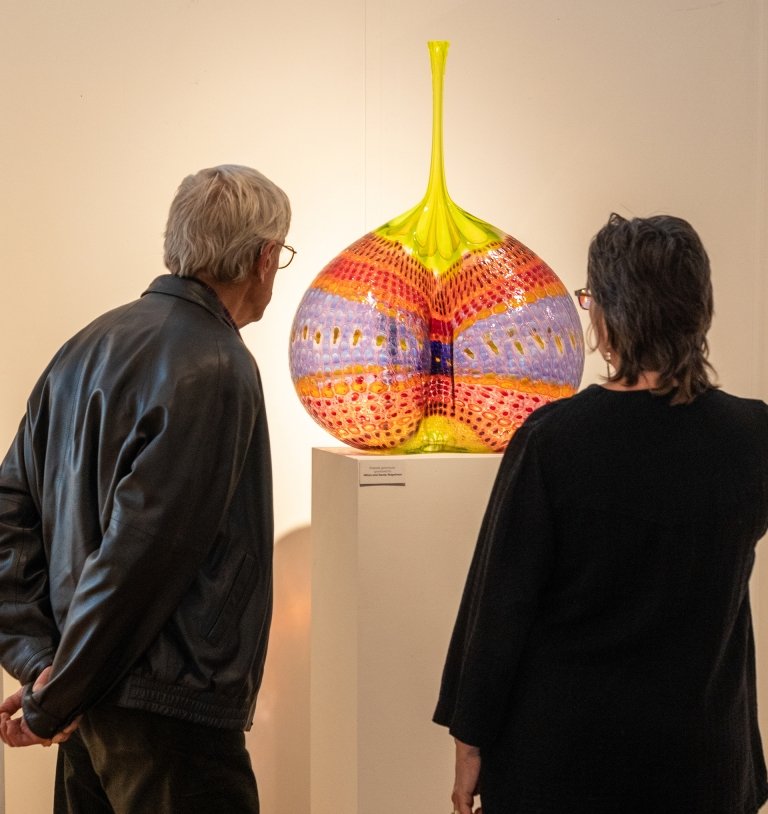 A man and a woman look closely at a brightly colored glass art vessel in display at a museum. 