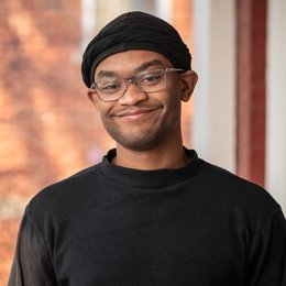 Man wearing black beanie hat and black sweater and glasses standing on front porch of ODI building