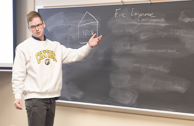 Derek Nafziger wears a Centre College sweatshirt as she stands at a black chalkboard teaching students about real estate insurance. 