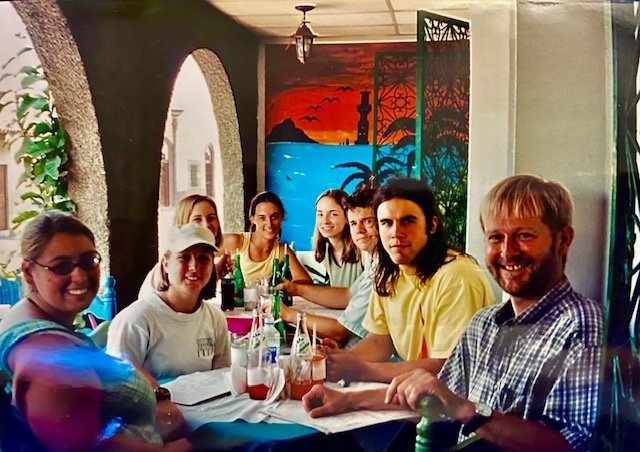 A group of men and women sit around a dining table in a brightly lit room. 