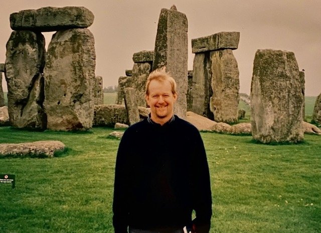 A man in a darm sweater stands in front of Stonehenge, an ancient collection of massive stones arranged in an intricate manor.