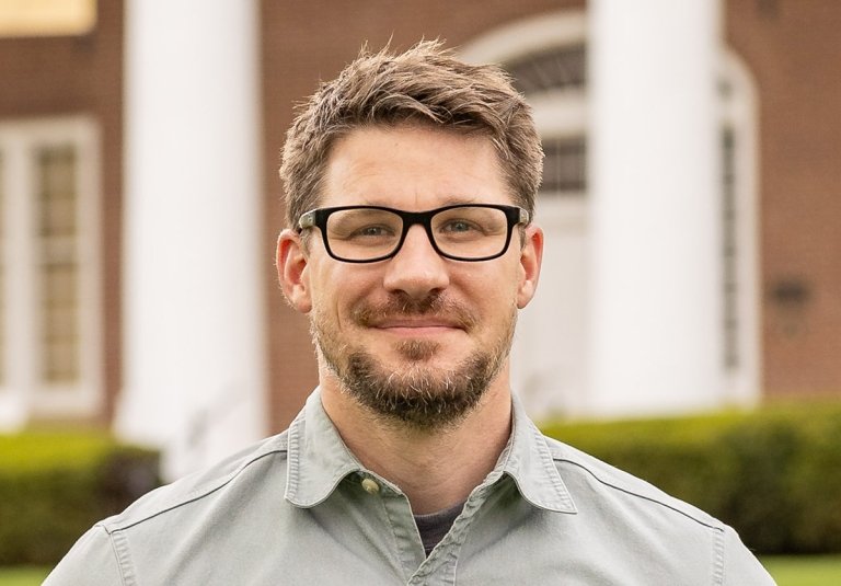 Man with dark hair with beard wearing glasses and grey button up dress shirt in front of old centre