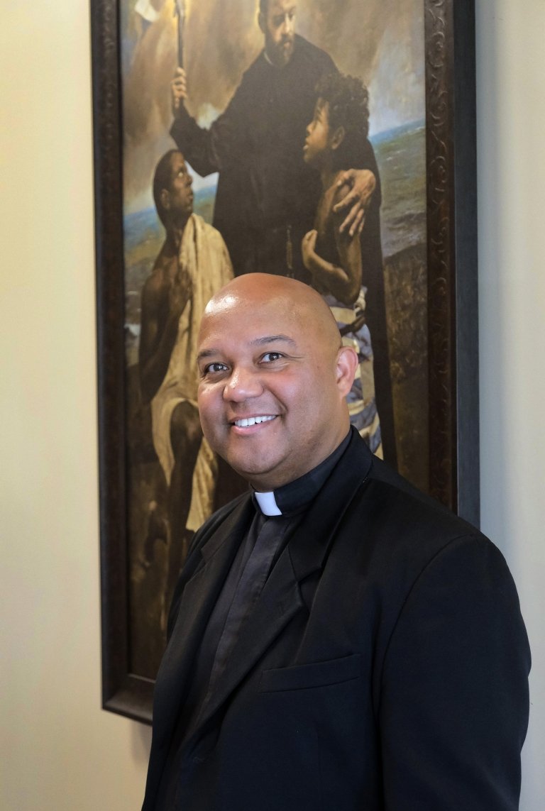 A man in a black priest's clothing and white collar poses in front of a painting.