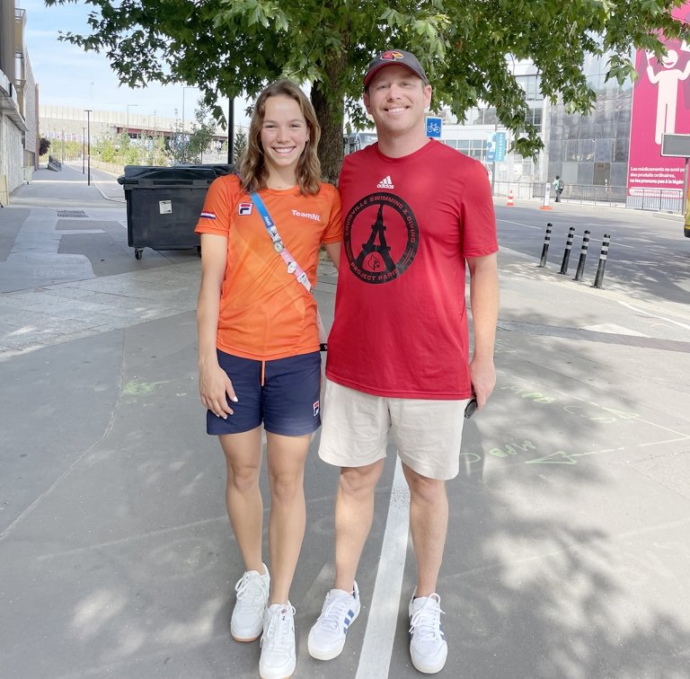 A woman and a man pose for a photo in front of a leafy green tree on a city sidewalk. 