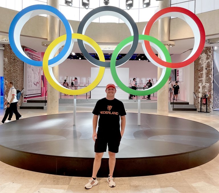 A man wearing a t-shirt and baseball cap poses in front of the Olympic Rings.
