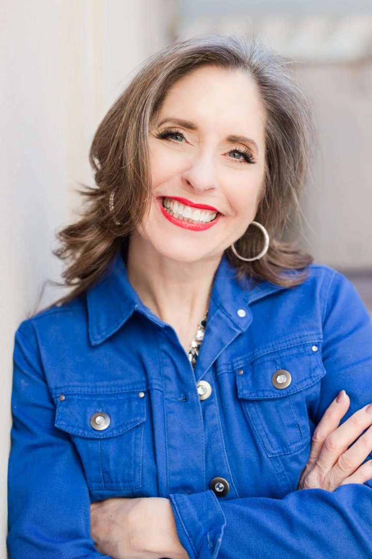 A woman poses for a photo, leaning against a white wall.